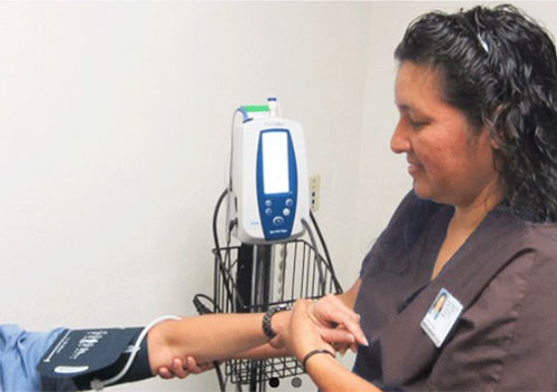 Image shows a health care person wearing brown scrubs administering a blood pressure check. Only the patient's arm is showing in this photo